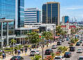Georgetown, Guyana city center with high-rise buildings, a busy palm-lined avenue, pedestrians, traffic, and the harbor with ships in the background