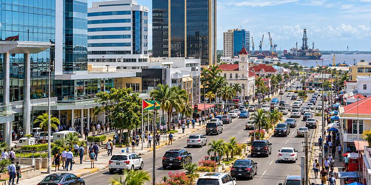Centre-ville de Georgetown, Guyana, avec des immeubles, une avenue animée bordée de palmiers, des piétons, de la circulation et le port avec des navires en arrière-plan