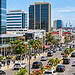 Georgetown, Guyana city center with high-rise buildings, a busy palm-lined avenue, pedestrians, traffic, and the harbor with ships in the background