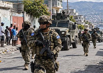 Des soldats armés de fusils patrouillent dans une rue de la ville aux côtés d’un véhicule blindé, tandis que des habitants observent depuis le bord de la route.