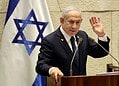 Israeli leader gestures while speaking at a podium, wearing a yellow ribbon, with an Israeli flag behind him
