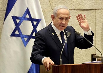 El líder israelí gesticula mientras habla en un podio, con una cinta amarilla y una bandera israelí detrás de él